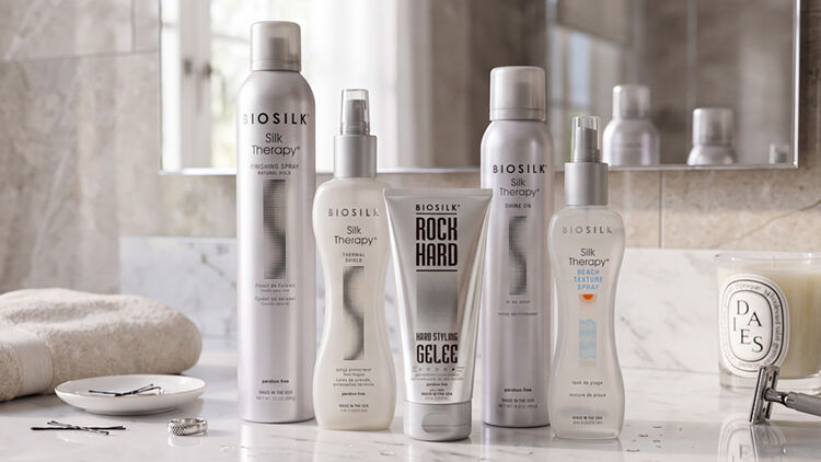 Five Biosilk styling products in silver and white, including hairsprays and Rock Hard gel&eacute;e, on a bathroom counter.